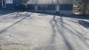 Un edificio pequeño de una sola planta, con fachada oscura y tejado inclinado. Se observa una puerta y una ventana pequeña en el lateral. El área circundante incluye un camino de entrada pavimentado y árboles que proyectan sombras.