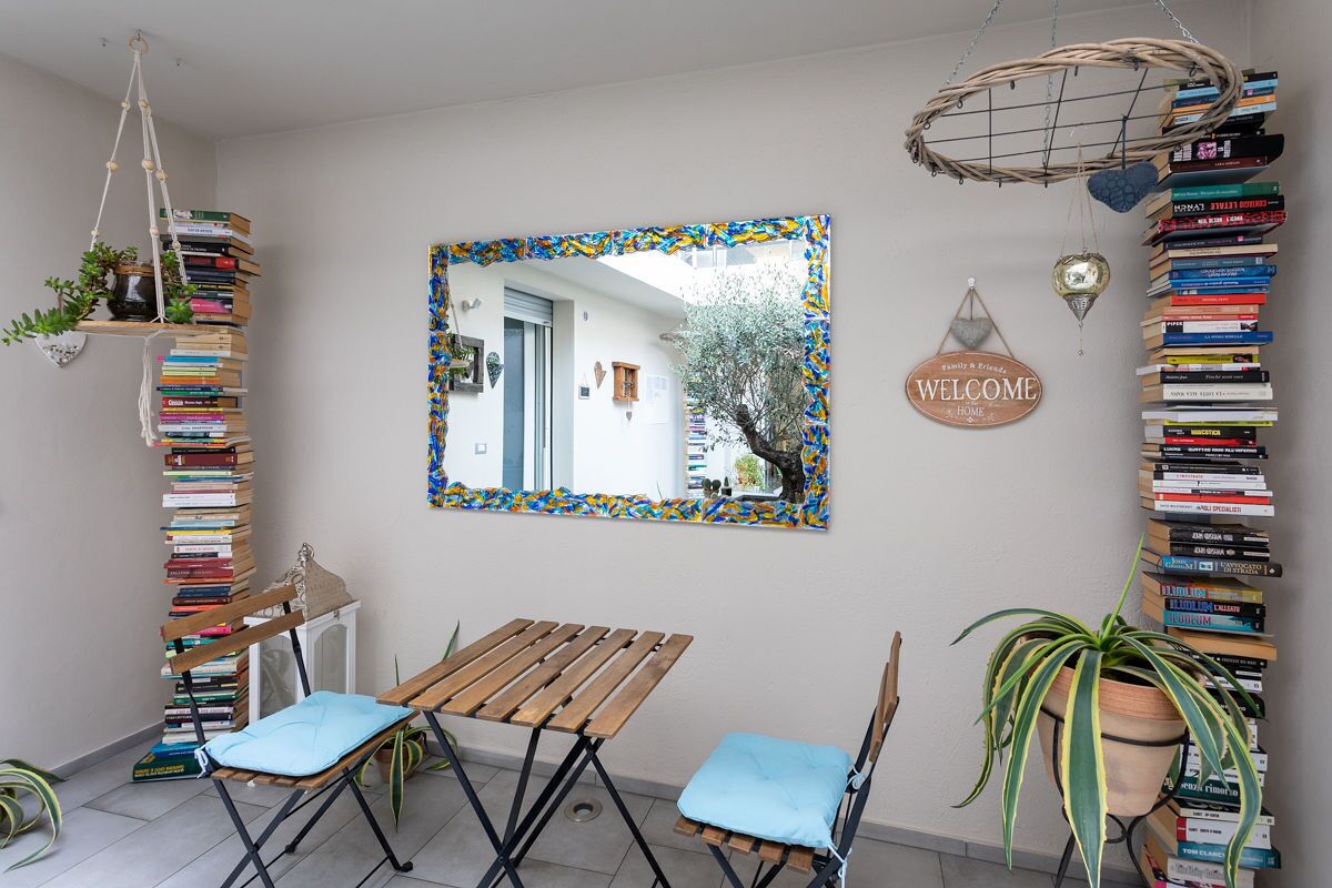 A room with a wooden table and two chairs, a mirror with a colorful frame, and a wall adorned with a 'WELCOME' sign and a bookshelf filled with books.