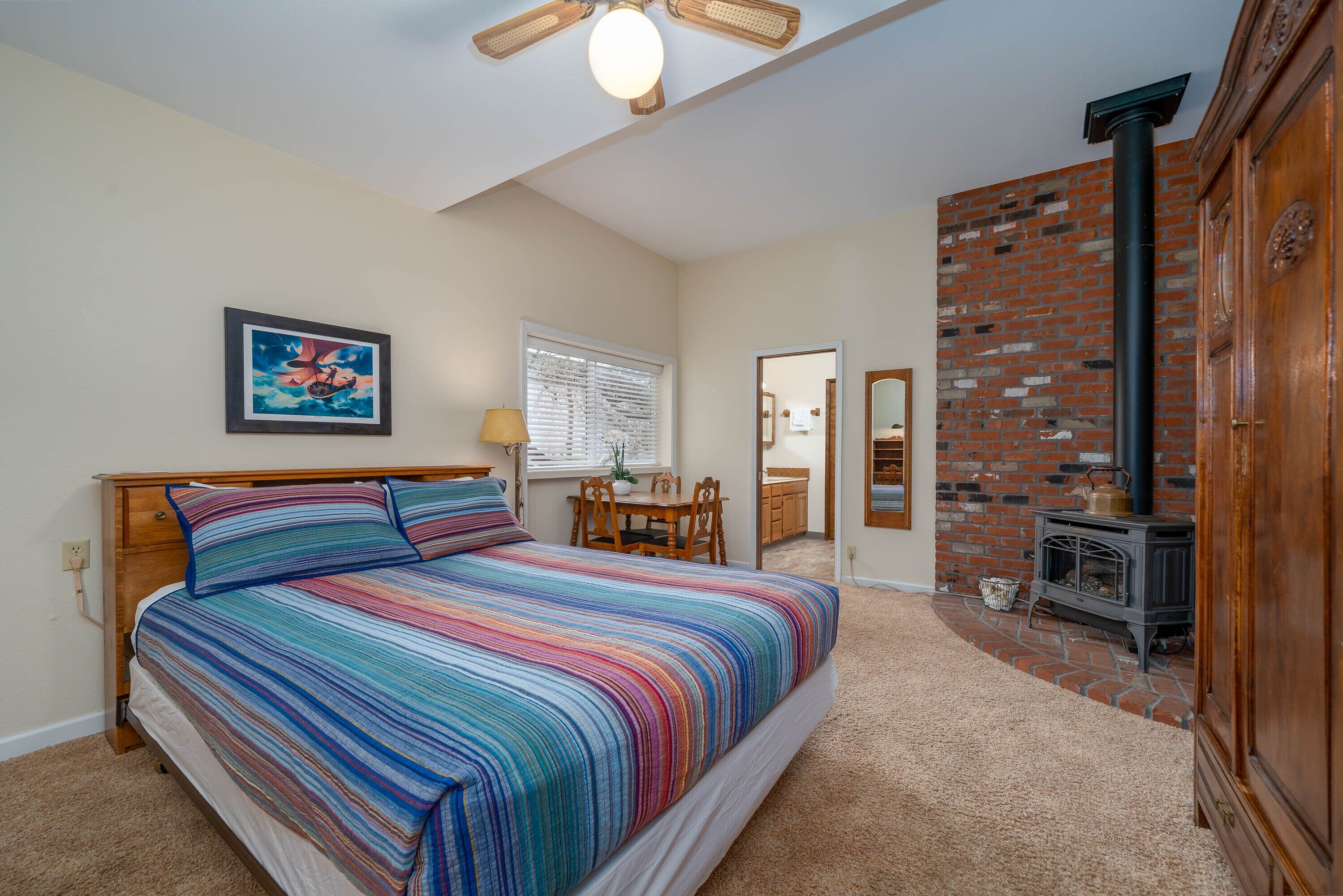 A bedroom with a large bed, a wooden headboard, a ceiling fan, a brick wall, a fireplace, and a dining area with a table and chairs.
