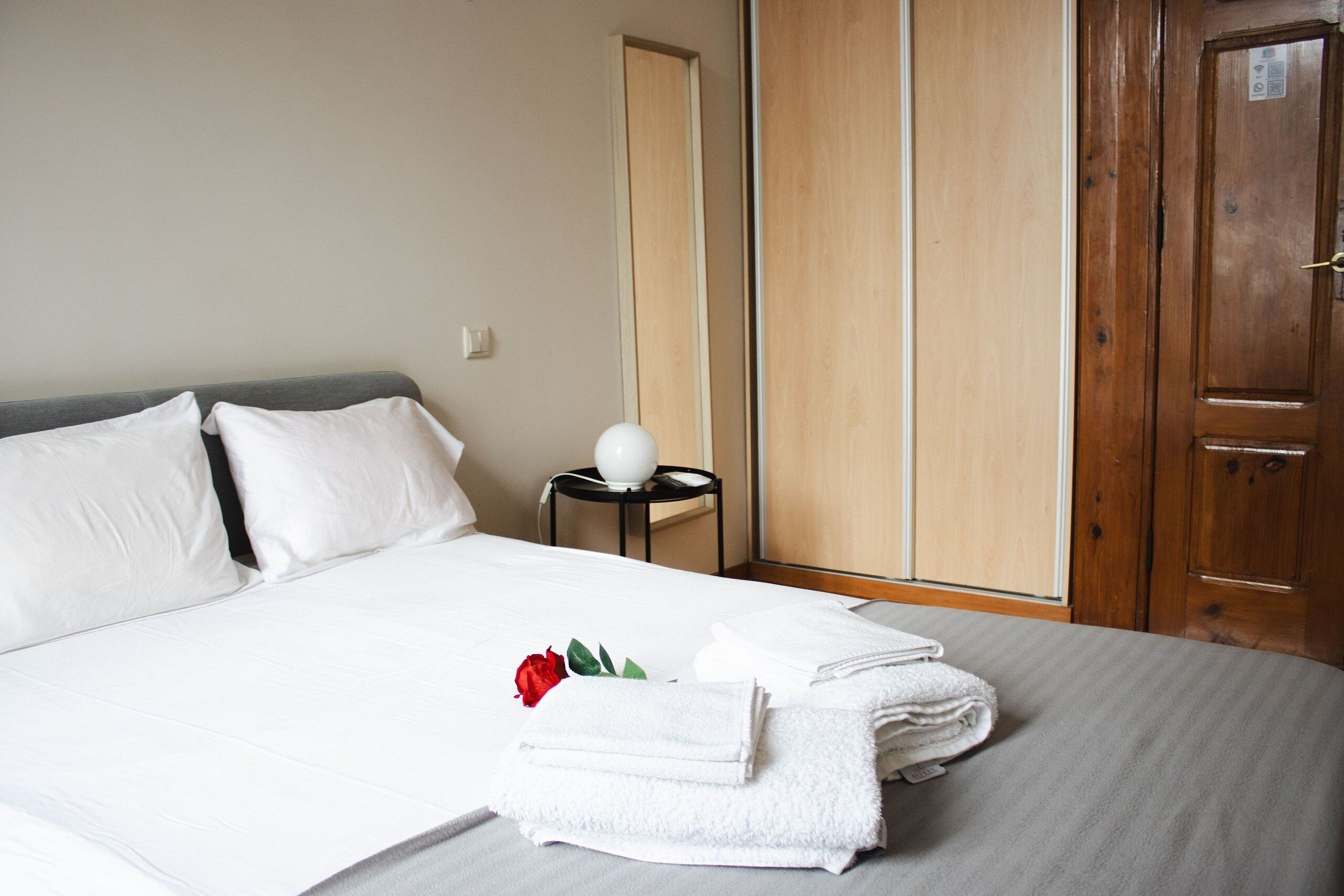 A neatly made bed with white linens and a red rose on a towel. There is a small round table with a white lamp beside the bed.