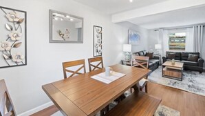 Un salon moderne avec une table à manger en bois, un canapé et une table basse. Les murs sont ornés d’œuvres d’art encadrées et un grand miroir est placé au-dessus de la table.