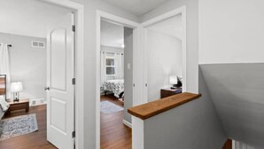Un couloir aux murs blancs et au plancher en bois mène à une chambre à coucher équipée d’un lit et d’une table de chevet.