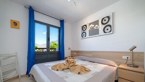 A neatly made bed with a patterned bedspread, a bedside table with a lamp, a wall-mounted artwork, and a window with blue curtains. - Casa LAGO (Playa Blanca)