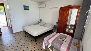 A small, clean room with a bed, a table with a patterned tablecloth, a wardrobe, and a wall-mounted air conditioner.