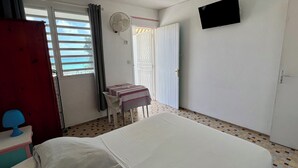 A room with a bed, a small table with a patterned tablecloth, a chair, a TV mounted on the wall, and a door leading to a balcony. - Studio Hibiscus n°2 with sea and mountain views, beach access, + cistern (Le Gosier)