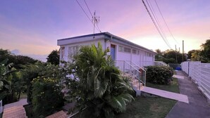 A single-story house with a white exterior, a small porch, and a staircase leading to the entrance. There is a palm tree in the foreground and a fence along the side of the property. - Studio Hibiscus n°2 with sea and mountain views, beach access, + cistern (Le Gosier)
