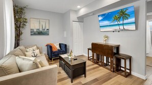 A living room with a sofa, coffee table, and a painting of a beach scene. - Wellington Garden (West Palm Beach)