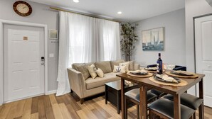 A modern living room with a beige sofa, a dining table set with placemats and a bottle, a clock on the wall, and a painting of sailboats. - Wellington Garden (West Palm Beach)