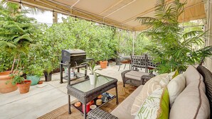 A covered patio area with wicker furniture, a glass-top table, and various potted plants. - Wellington Garden (West Palm Beach)
