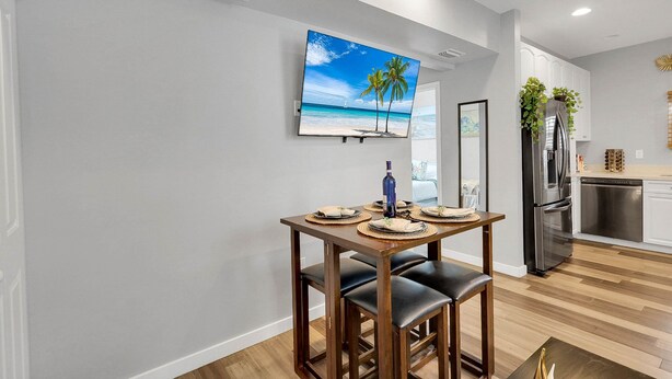 A modern kitchen with a stainless steel refrigerator, a wooden dining table with chairs, and a wall-mounted flat-screen TV displaying a beach scene. - Wellington Garden (West Palm Beach)