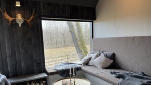A cozy room with a large window, a wooden wall, a grey sofa, a round table with candles, and a fireplace.
