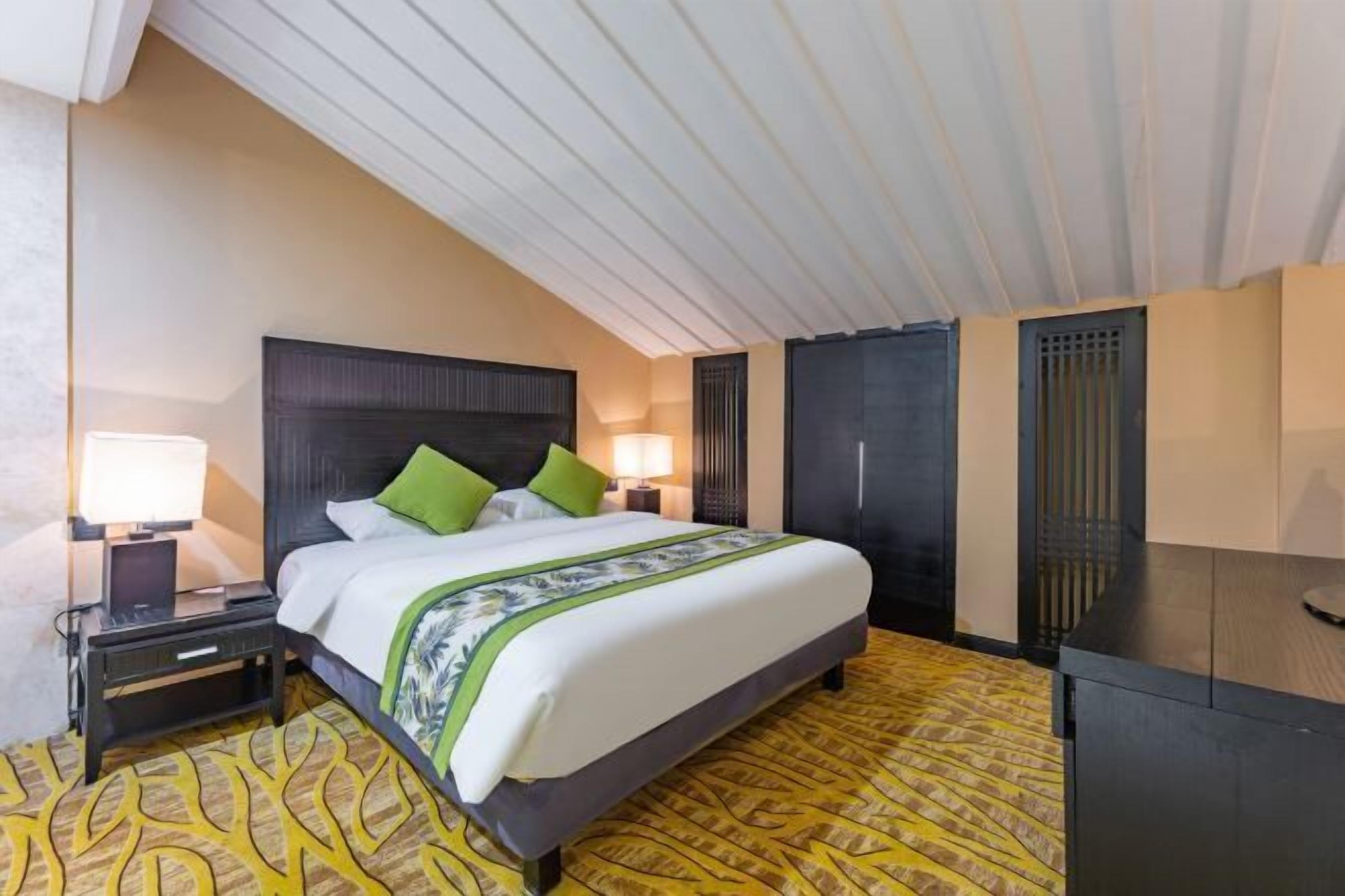 A hotel room with a bed, bedside tables, lamps, a desk, and a patterned carpet.