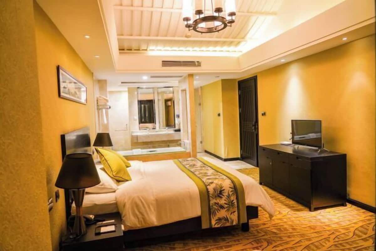 A hotel room with a bed, a desk with a TV, a mirror, and a chandelier.