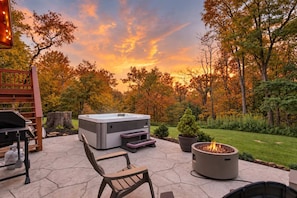 A hot tub, outdoor seating, and a fire pit in a garden with trees and a sunset. - The Juniper Cabin | Cozy Hideaway w/ Hot Tub, Screened Porch + Fire Pit (Linden)