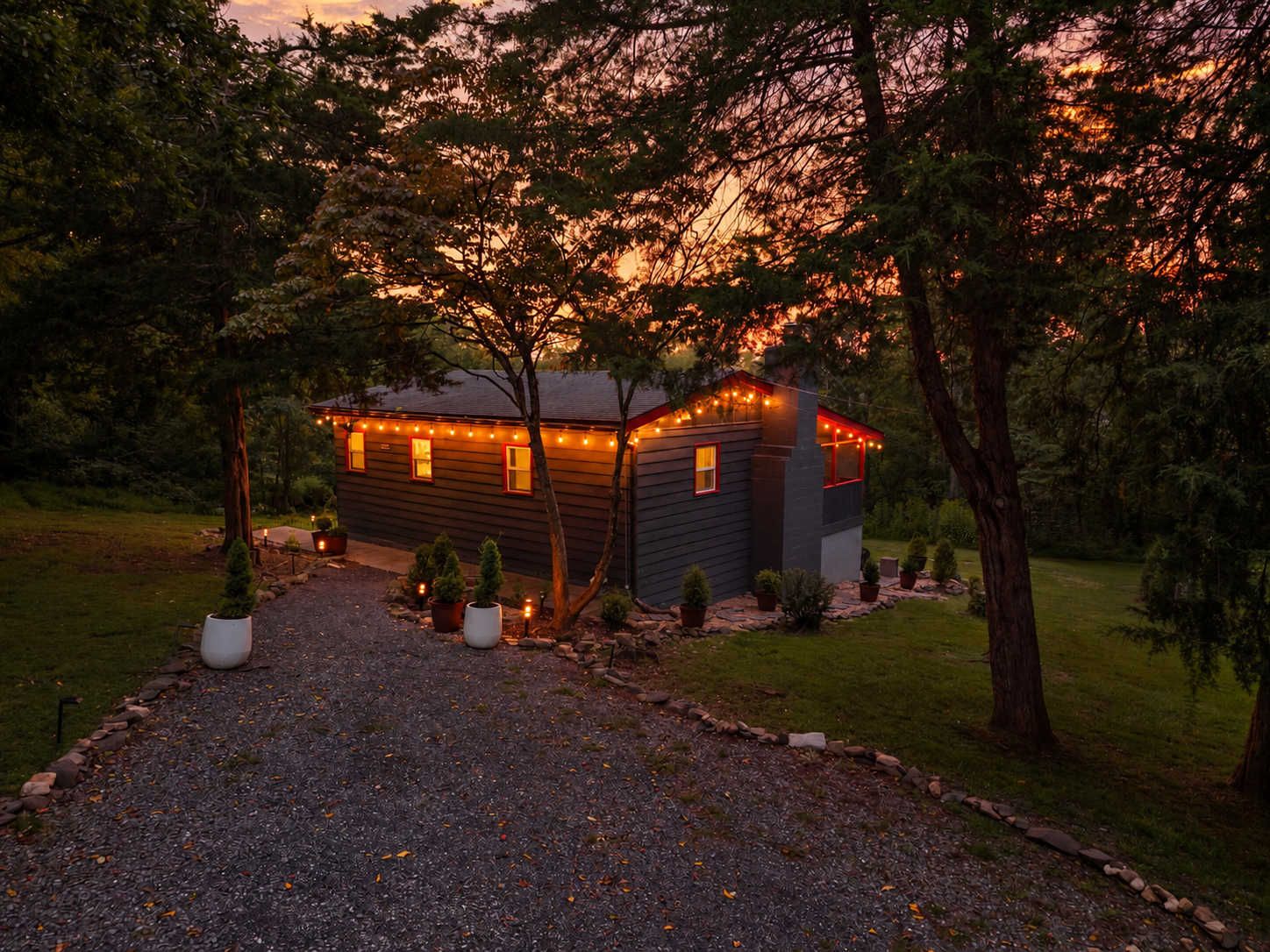 A tree-lined gravel driveway leads to the cabin, lit by string lights along the porch.