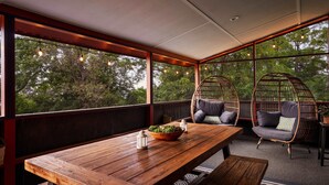 A wooden table with a bowl of plants, two wicker chairs with cushions, and a view of trees through large windows.