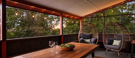 A wooden table with two chairs and a bowl of fruit, set against a backdrop of trees and a view through large windows.