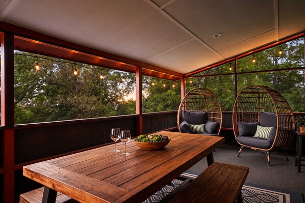A wooden table with two chairs and a bowl of fruit, set against a backdrop of trees and a view through large windows.