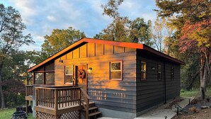 A small cabin with a wooden deck and a front porch, surrounded by trees and a grassy area.