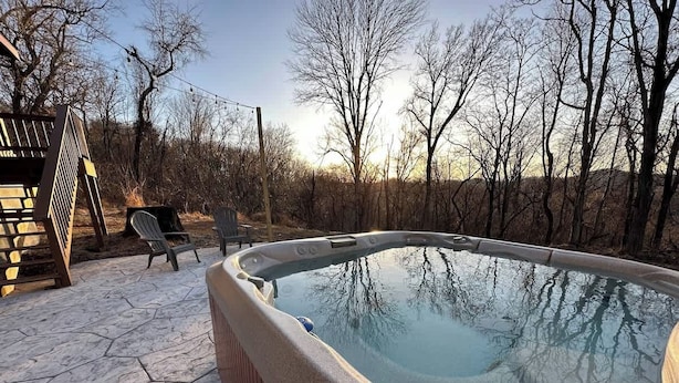 A hot tub with a stone patio, surrounded by trees and a wooden deck.