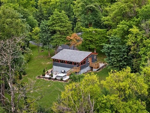 A cabin surrounded by dense forest, with a deck and outdoor seating area. - The Juniper Cabin | Cozy Hideaway w/ Hot Tub, Screened Porch + Fire Pit (Linden)