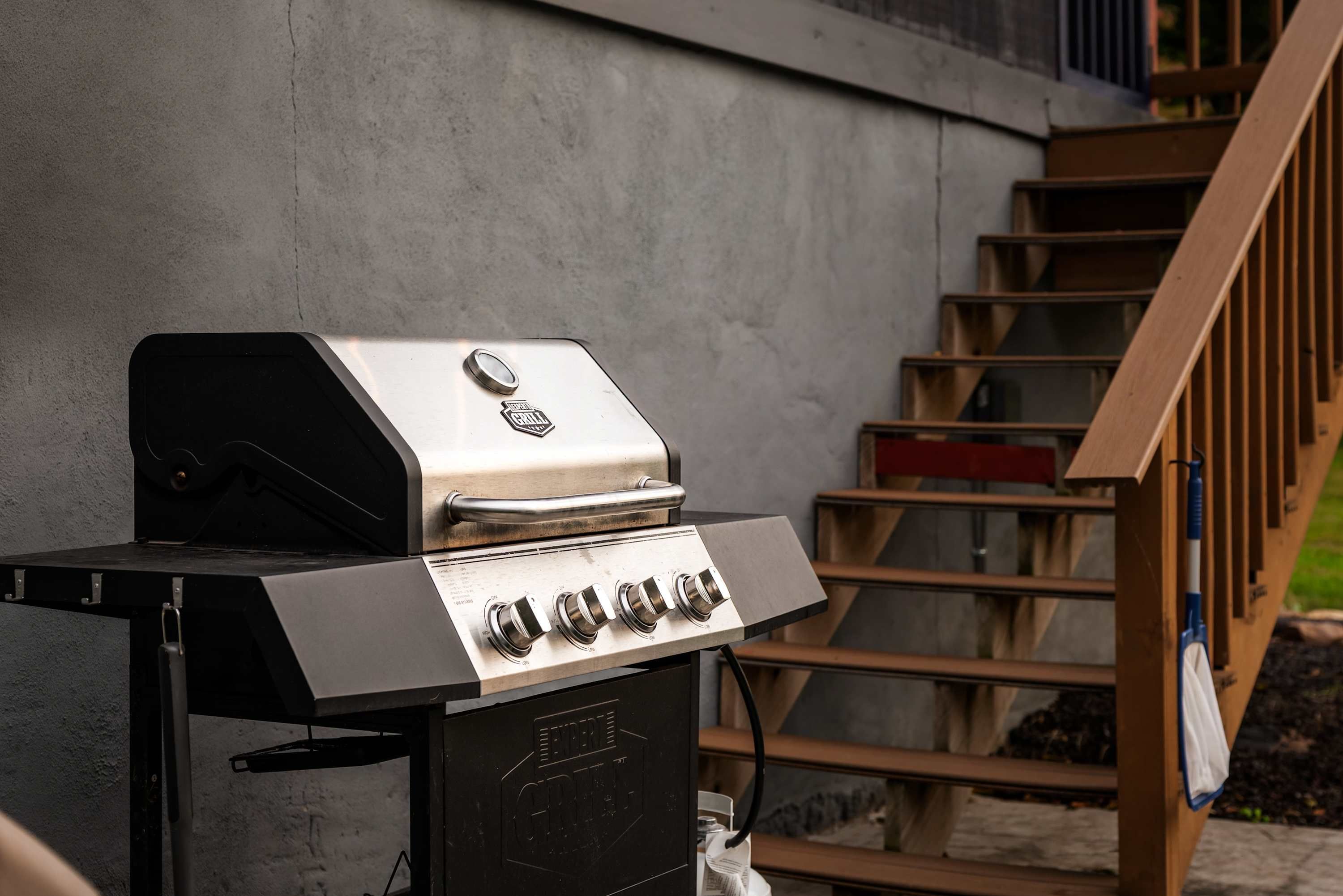 A stainless steel barbecue grill with four burners, positioned against a concrete wall with a wooden staircase in the background.