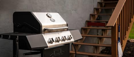 Una parrilla para barbacoa de acero inoxidable con cuatro quemadores, ubicada frente a una pared de concreto y una escalera de madera al fondo.
