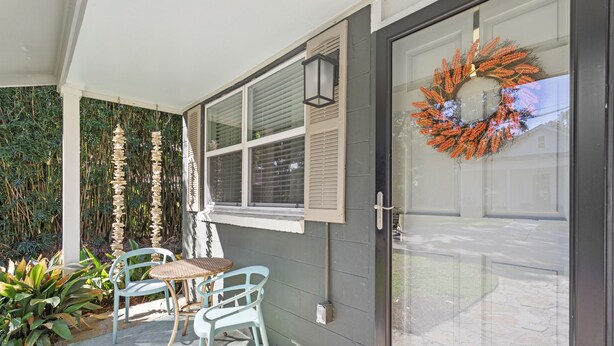 A covered patio with a table and chairs, a decorative wreath on the door, and a view of the house through the window. - Downtown Cottage! Walk/Cart Everywhere, 2 King Beds (Ocean Springs)
