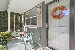 A covered patio with a table and chairs, a decorative wreath on the door, and a view of the house through the window. - Downtown Cottage! Walk/Cart Everywhere, 2 King Beds (Ocean Springs)