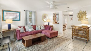 A living room with a sectional sofa, a coffee table, a dining area with a wicker chair, and a ceiling fan. - Downtown Cottage! Walk/Cart Everywhere, 2 King Beds (Ocean Springs)