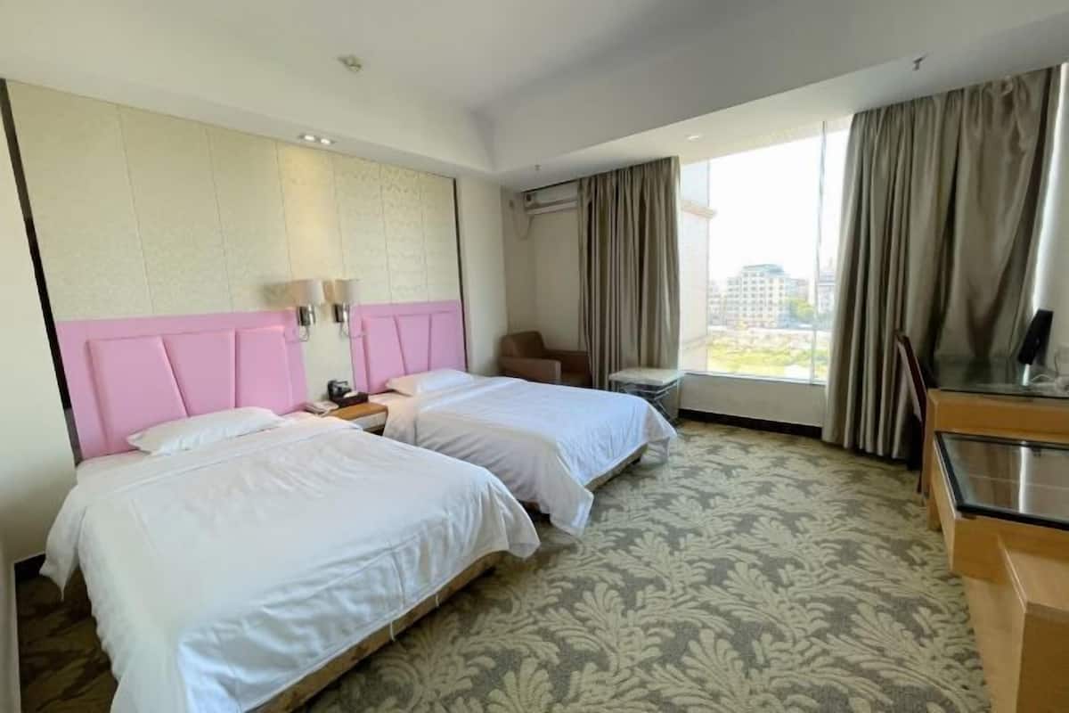 A hotel room with two beds, a desk, a chair, and a view of buildings.