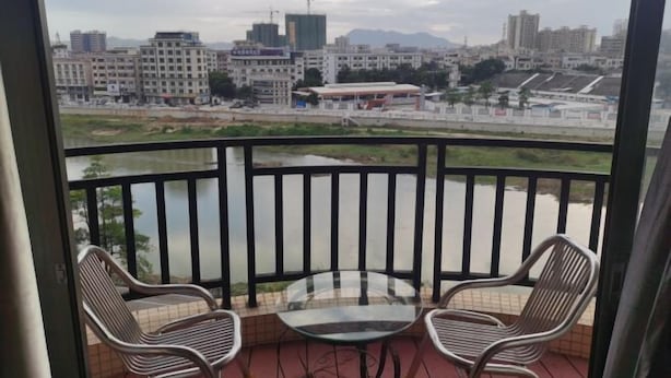 A balcony with two wicker chairs and a glass table, overlooking a river and a cityscape with buildings and construction. - Jufu Hotel (Enping)