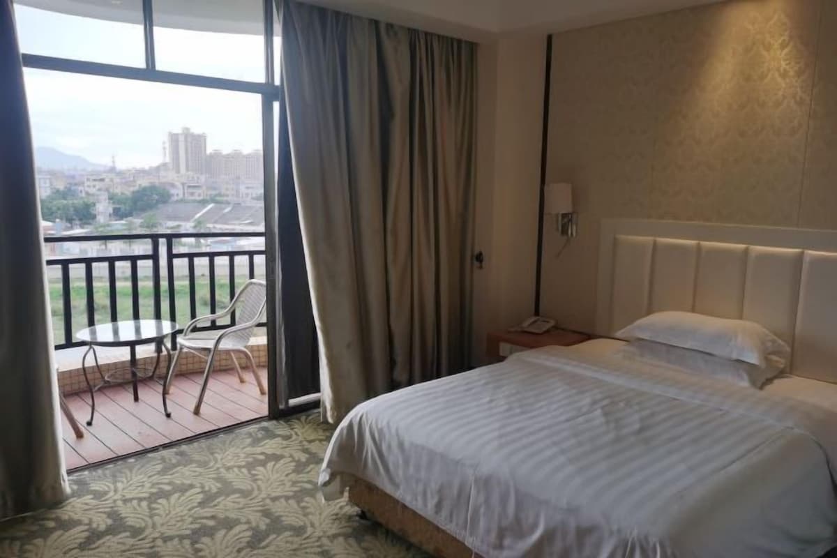 A hotel room with a large bed, a nightstand, a chair, and a balcony with a view of the city.