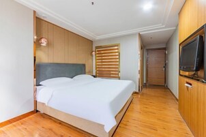 Un dormitorio con cabecera de madera, cama con ropa de cama blanca, televisor sobre un mueble de madera y suelo de madera.