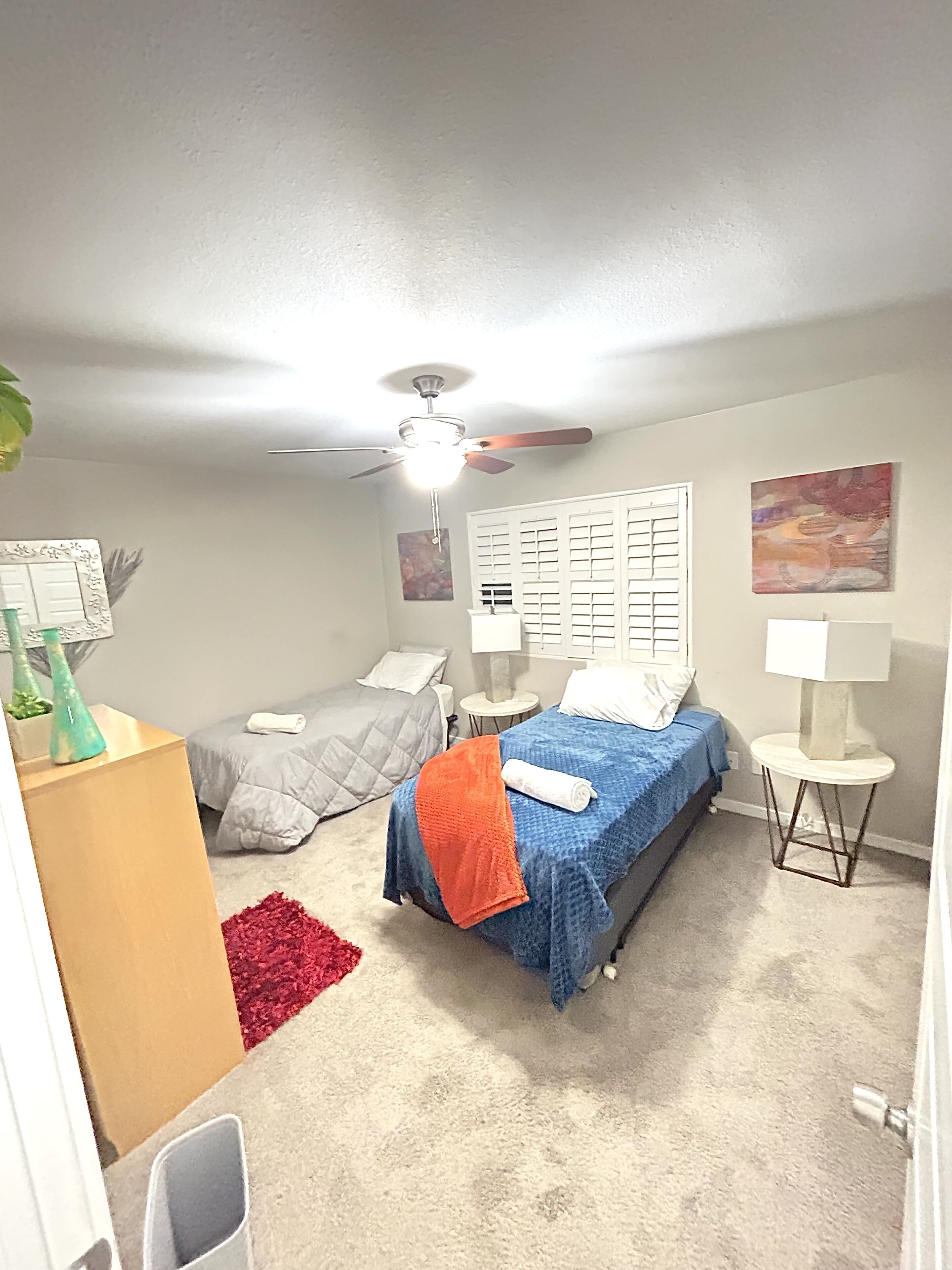 A bedroom with two beds, a ceiling fan, a red rug, and a small table with a lamp.