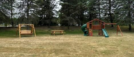 Une aire de jeux avec des balançoires, un toboggan et une petite structure en bois, le tout situé dans un espace verdoyant, avec des arbres en arrière-plan.