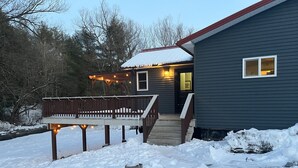 Una casa con terraza de madera, un porche adornado con luces y un patio cubierto de nieve.