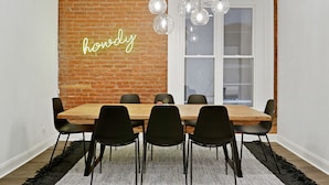 Een eethoek met een bakstenen muur, een neonbord, een houten tafel, zwarte stoelen en hangende bolvormige lampen.