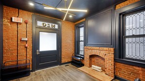 Een kamer met een bakstenen muur, een zwarte deur met een 'EXIT'-bordje, een open haard en een raam met een metalen rooster.