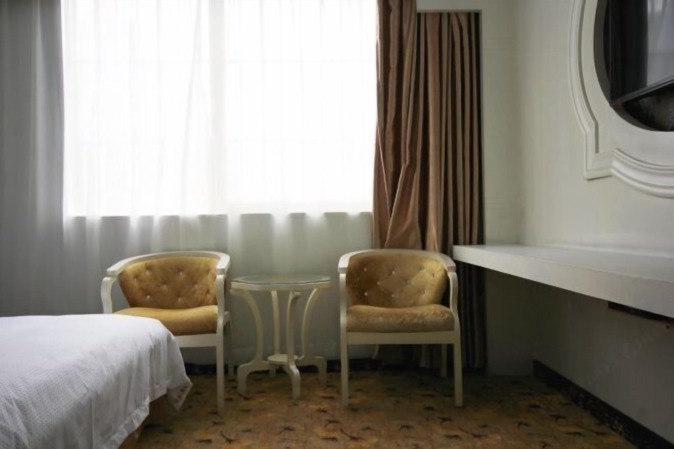 A hotel room with a bed, two armchairs, a small round table, a window with curtains, and a white wall with a round mirror.