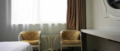 A hotel room with a bed, two armchairs, a small round table, a window with curtains, and a white wall with a round mirror.