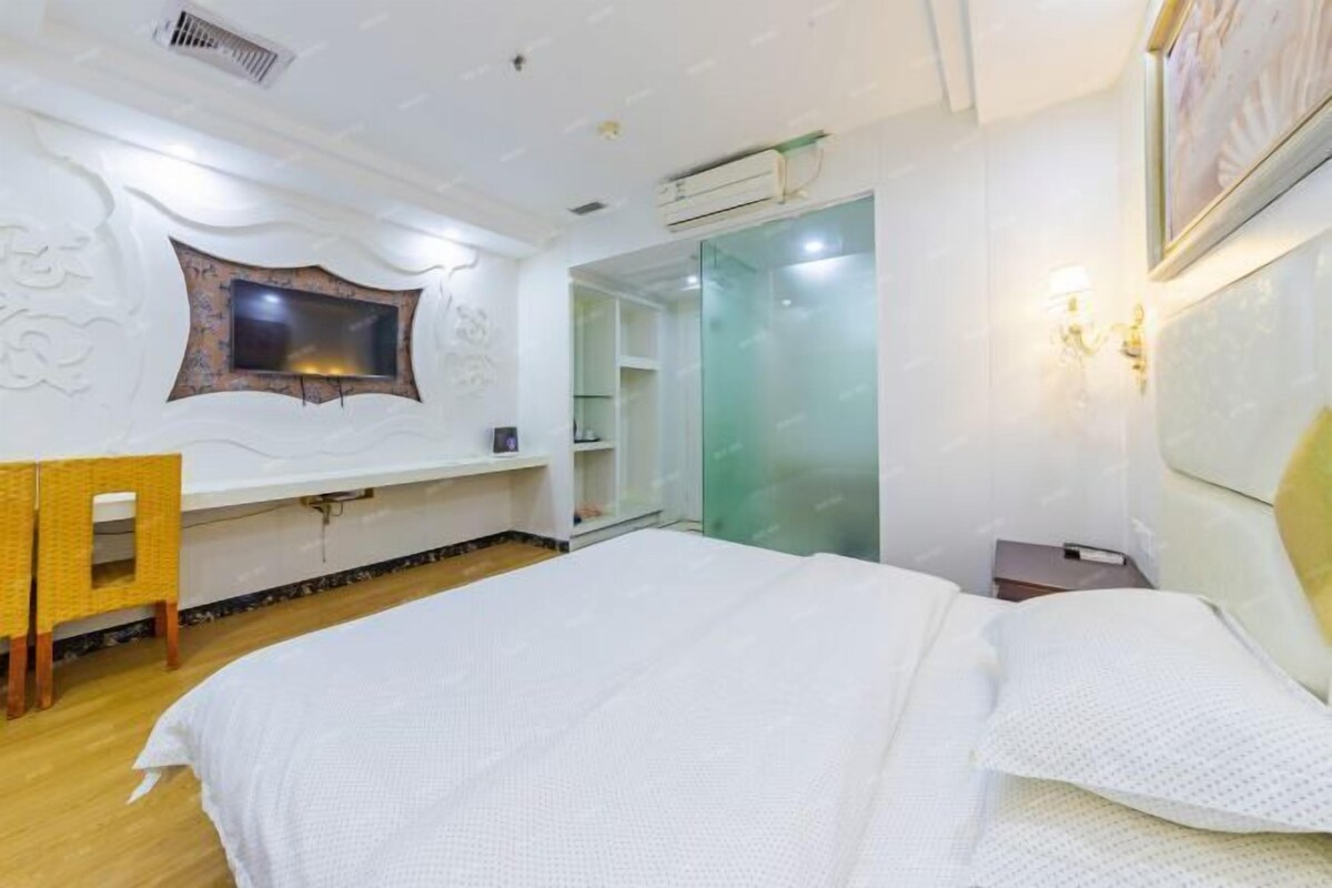 A hotel room with a large bed, a small desk, a chair, a wardrobe, and a glass door leading to a bathroom.