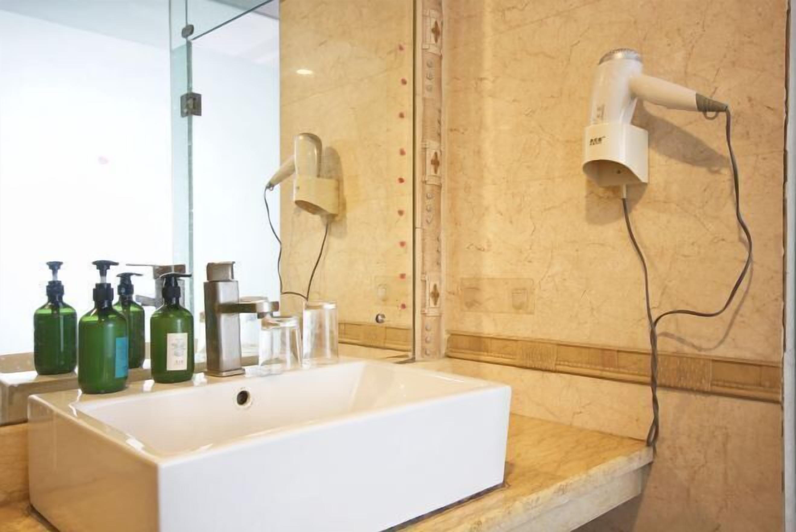 Een badkamer met een groot wit wastafel, groene handzeepflessen, een spiegel en twee haardrogers aan de muur.