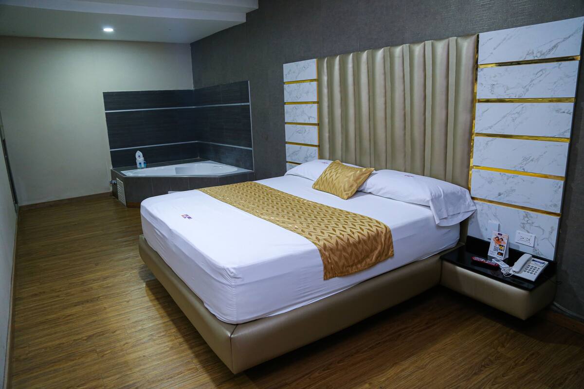 A hotel room with a large bed, a bedside table with a phone and a remote control, a small bedside table with a magazine, and a bathroom with a bathtub and a mirror.