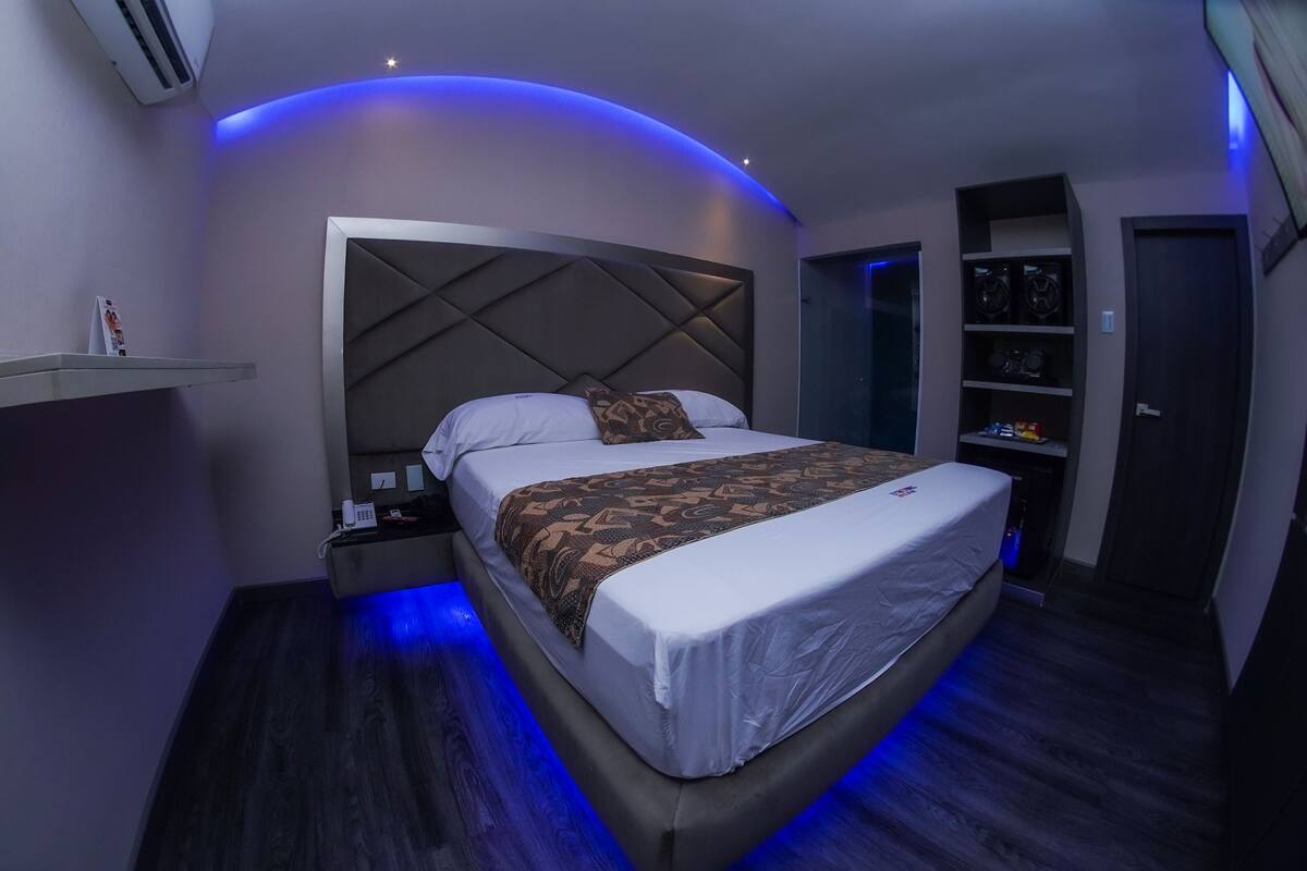 A modern hotel room with a large bed, a headboard with a geometric pattern, a bedside table with a phone and a magazine, and a built-in shelf with speakers.