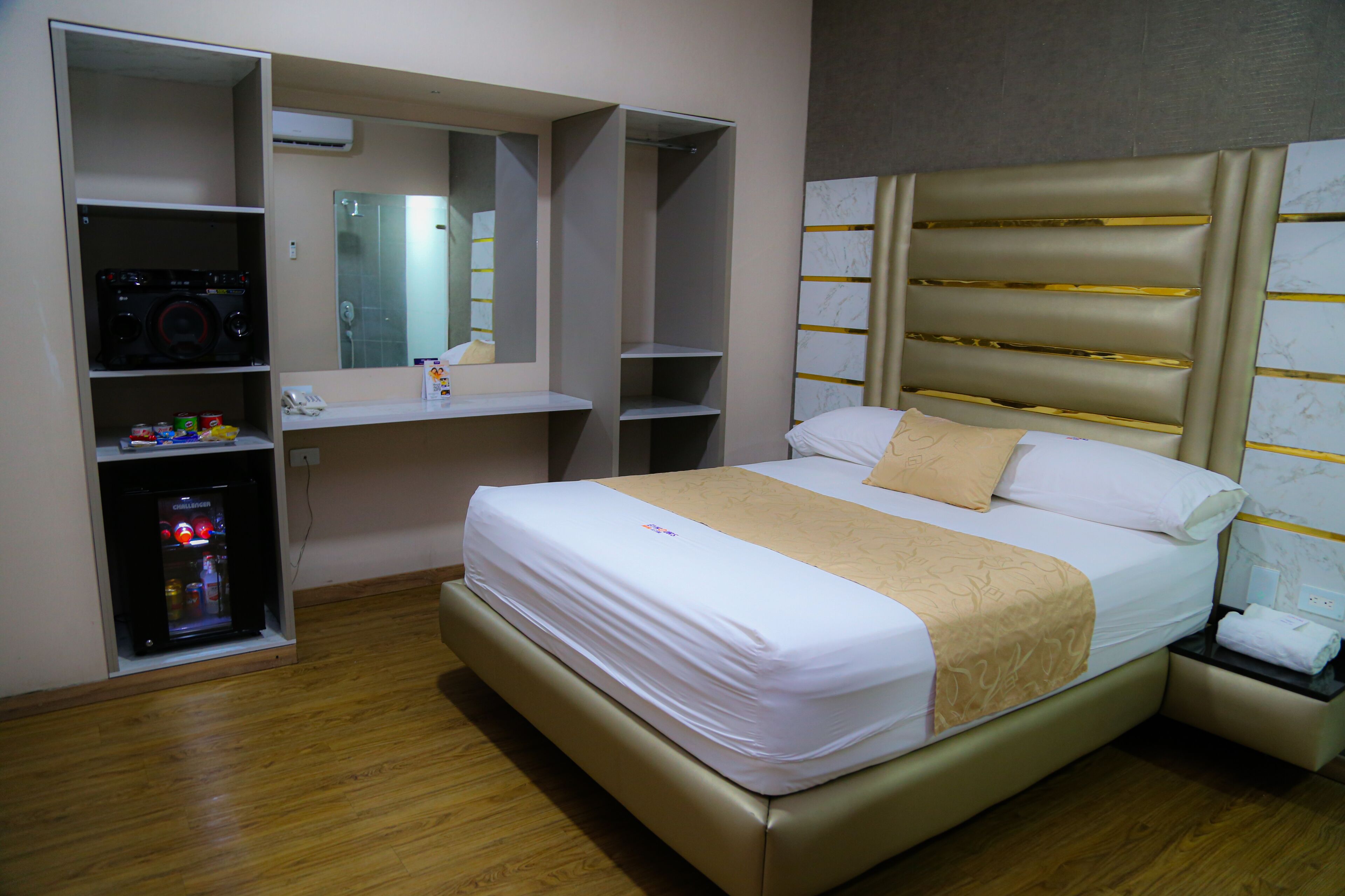 Habitación de hotel con cama, minibar, espejo y estantería empotrada.
