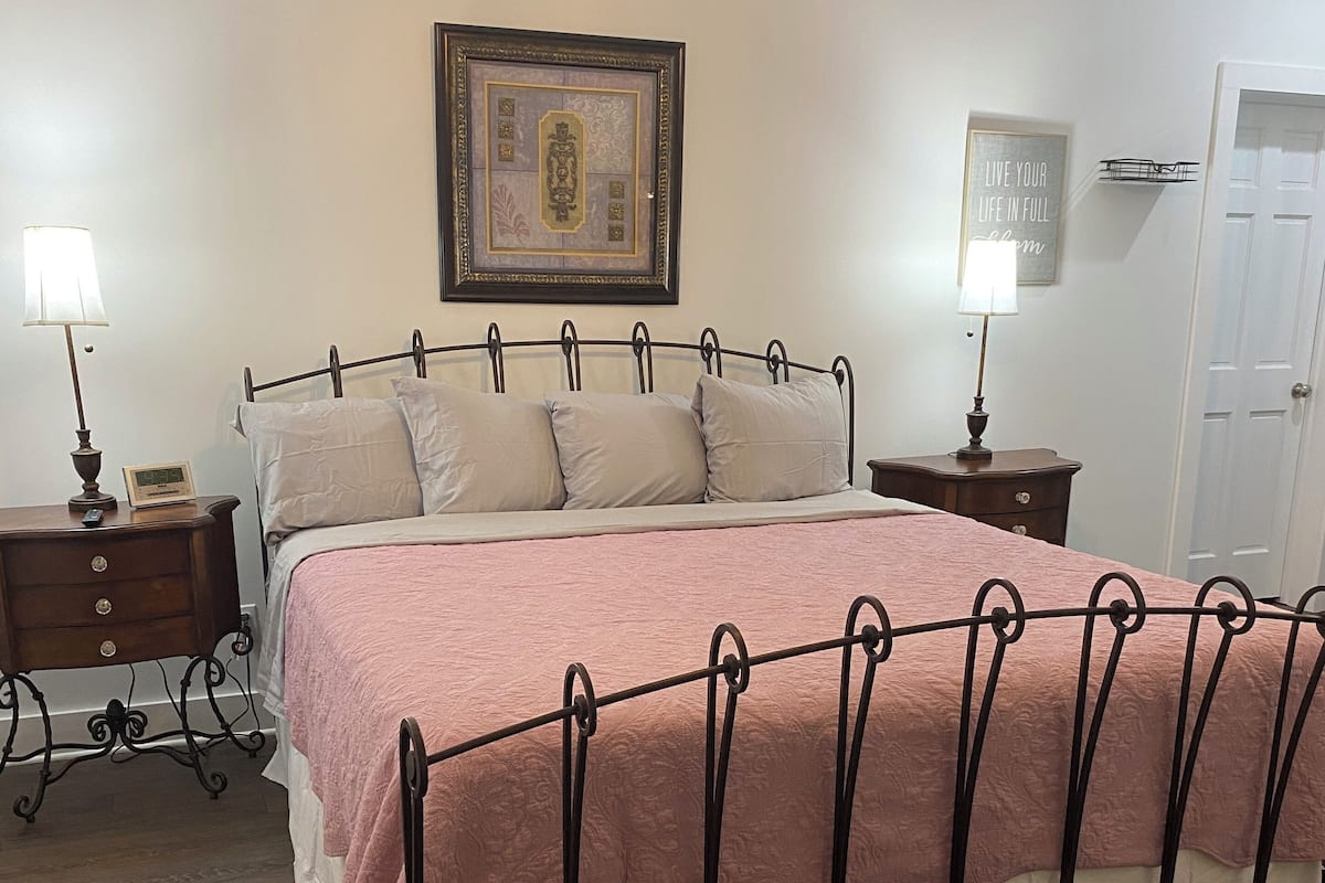 A neatly arranged bedroom with a metal bed frame, a pink bedspread, and a wooden floor. There are two bedside tables with lamps, a framed picture on the wall, and a door in the background.