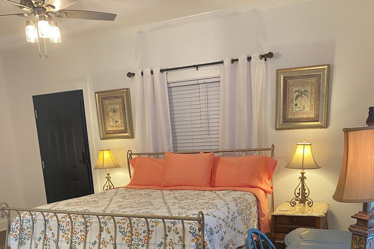 A bedroom with a bed, a desk, a chair, a ceiling fan, two bedside lamps, a window with curtains, and a door.