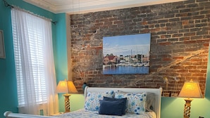 A bedroom with a white bed, a brick wall, and a painting of a harbor. - Little Inn on Main (Washington)