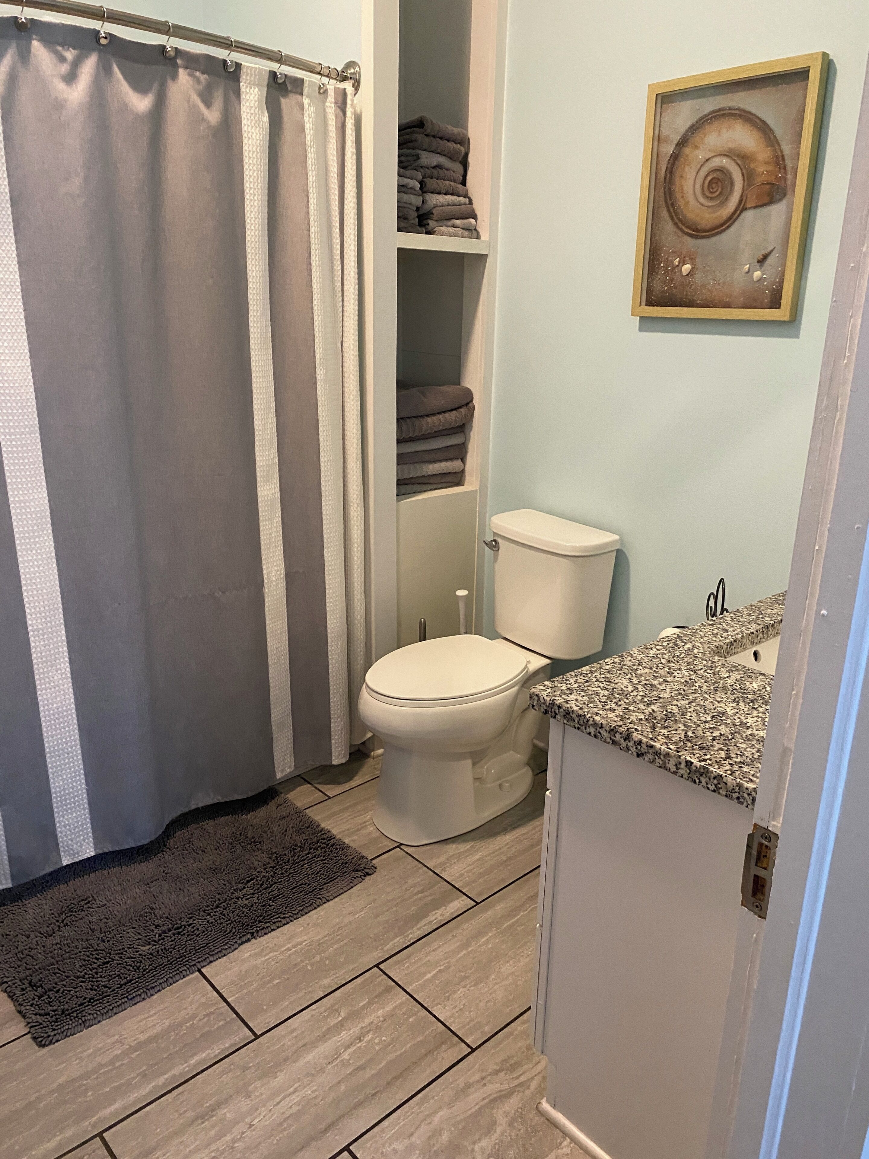 A bathroom with a toilet, a shower curtain, a towel rack with folded towels, and a framed picture on the wall.